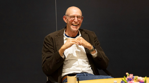 Michael Pollan, laughing, sitting at a table while lecturing
