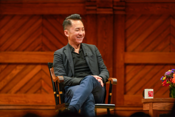 Viet Thanh Nguyen, seated with his legs crossed. He is wearing a black shirt, dark grey jacket, and blue jeans.
