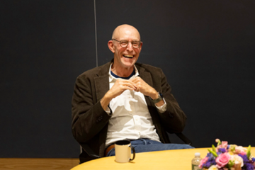 Michael Pollan, laughing, sitting at a table while lecturing
