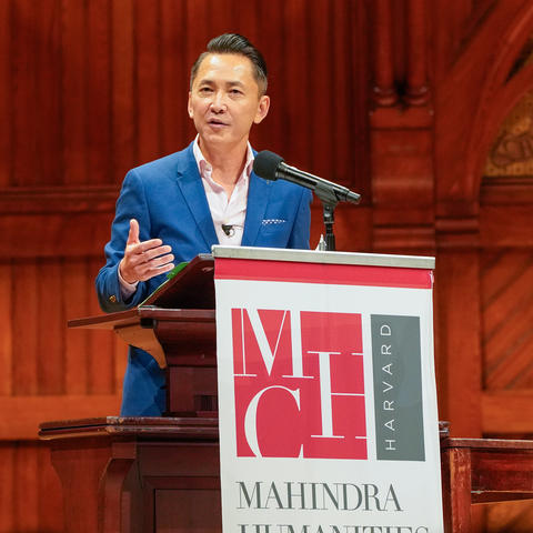 Viet Thanh Nguyen, in a blue suit, speaking behind a podium with the Mahindra Humanities Center Logo on the front.